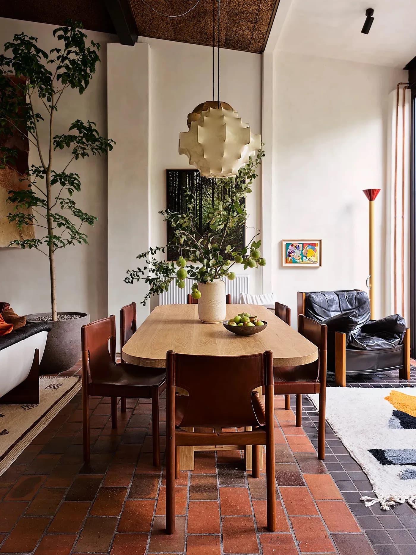 Dining room of singer Troye Sivan’s home. The back of the sofa, end of the rug, and large potted tree on the left define the end of the living room. Just to the right, a column marks the start of the dining area, where we can see the centered dining room table with potted plant in its center and pendant lamp above it. This creates a strong focal point and sense of balance.